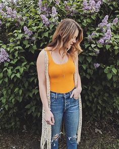 a woman standing in front of purple flowers wearing ripped jeans and an orange tank top