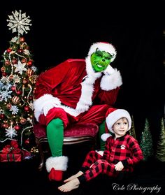 a little boy sitting in front of a christmas tree next to a man dressed as the grin