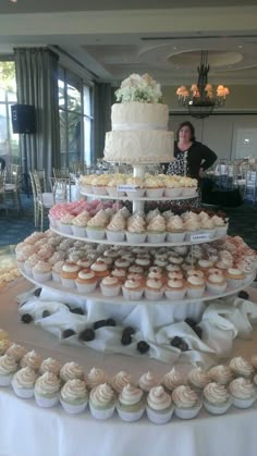 a woman standing next to a large tiered cake and cupcakes on a table