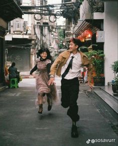 a man and woman running down the street in an alleyway with buildings on either side