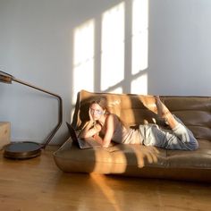 a woman laying on top of a brown couch next to a lamp and a laptop