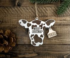 a cow ornament hanging on a wooden wall next to pine cones and evergreen needles