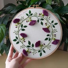 a hand is holding up a embroidery hoop with purple flowers and green leaves on it