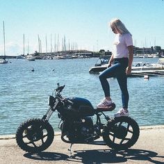 a woman standing on the back of a motorcycle next to a body of water with boats in the background