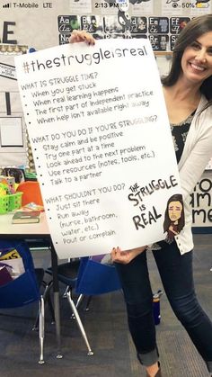 a woman holding up a sign in front of a classroom desk with posters on it