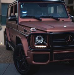 a brown mercedes g - class is parked in front of a building with its lights on