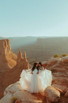 two women in wedding dresses standing on rocks at the edge of a cliff overlooking canyon