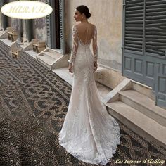 a woman standing in front of a building wearing a wedding dress with long sleeves and an open back