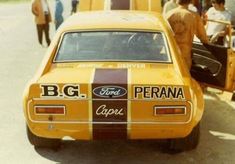 an old yellow car is parked on the side of the road as people walk by