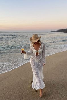 a woman in a white dress and hat walking on the beach holding a glass of wine