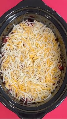 a crock pot filled with shredded cheese on top of a pink table next to utensils