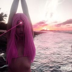 a woman with pink hair standing in front of the ocean holding her arms above her head