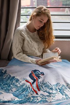 a woman reading a book while sitting on a bed with a mountain scene printed on it