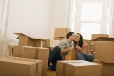 a man and woman sitting on cardboard boxes in the middle of a room with windows