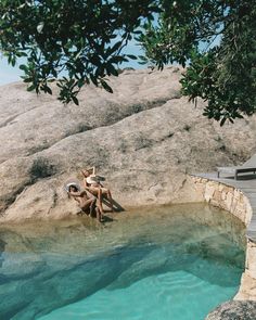 two people sitting on the edge of a pool in front of a large rock formation