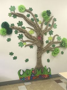 a tree made out of paper with green leaves and flowers on the branches, sitting in front of a white wall