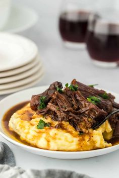 a white plate topped with mashed potatoes covered in beef and gravy next to wine glasses