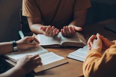 three people sitting at a table with open books in front of them and one person holding out their hands