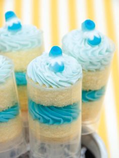 four cupcakes with blue frosting in plastic cups on a tray next to a yellow and white striped tablecloth