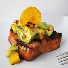 a piece of meat with avocado and sliced pineapple on top, sitting on a white plate next to a fork