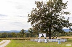 an outdoor ceremony set up with white chairs under a large tree and mountains in the background