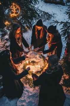 four people standing around a christmas wreath with candles in the snow at night, surrounded by evergreens