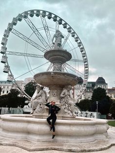 a woman standing in front of a water fountain with a ferris wheel behind her on a cloudy day