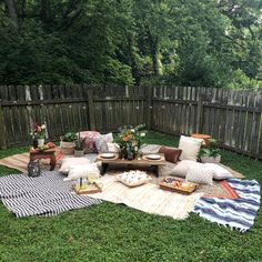 an outdoor seating area with blankets, pillows and flowers on the grass in front of a wooden fence