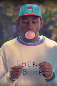 a man wearing a white shirt and blue hat blowing bubble gum on his tongue while standing in front of trees