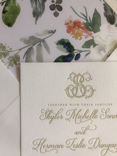 an elegant wedding card with gold lettering and floral designs on it, sitting next to a white envelope