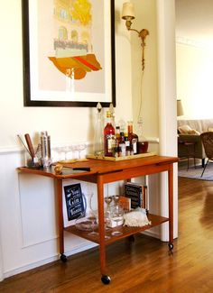 a bar cart with liquor bottles and glasses on it in front of a framed painting