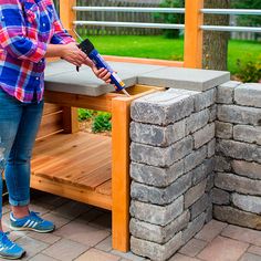 a woman sanding on top of a wooden bench next to a stone wall with a hammer