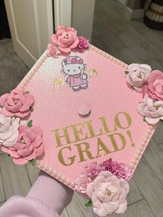 a pink graduation cap with hello kitty on it and flowers around the top that says hello grad