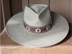 The Bandit is a stand out hat that captures the feeling of the American West. . The base of the hatband is distressed leather that is then layered with imprinted brown leather, bolo sliders and lots of nailspots. It is finished with this colorful feather combo. The brim of the LID custom hat body is 4" in diameter and the crown is 4.5". This is a stiff hat. Available in Putty or Pecan All hats are made to order. Allow up to 4 weeks for your hat to ship. Leather High Crown Hat Bands For Rodeo, Rustic Brown Hat With Flat Crown, Adjustable Distressed Hats For Country Events, Distressed Adjustable Hats For Country Events, Adjustable Distressed Fedora With Flat Brim, Brown Country Hat With Flat Crown, Western Style Brown Hat With Flat Crown, Country Style Brown Hat With Flat Crown, Rustic Ranch Hat With Flat Crown