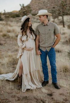 a pregnant woman in a long dress and cowboy hat standing next to a man with his arm around her waist