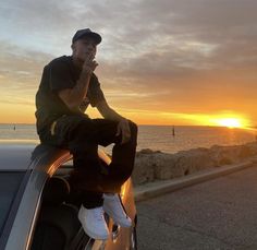 a man sitting on the hood of a car in front of an ocean at sunset