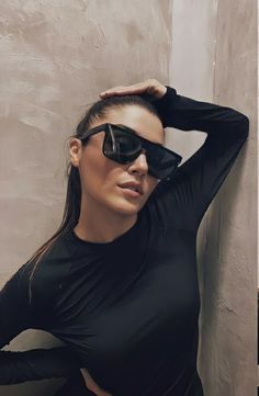 a woman in black shirt and sunglasses standing next to a wall with her hands on her head