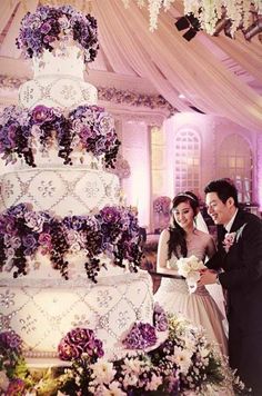 a bride and groom standing next to a wedding cake with purple flowers on the side