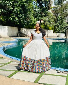 a woman standing in front of a pool wearing a white dress with multicolored squares on it