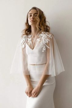 a woman wearing a white wedding dress with sheer sleeves and flowers on the shoulders, standing in front of a white wall