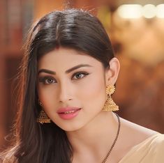 a woman with long dark hair wearing gold earrings and a beige dress is looking at the camera