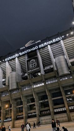 people are walking around in front of a stadium building at night with the lights on