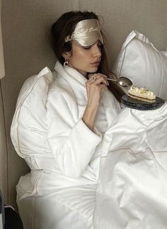 a woman laying in bed with a blindfold on her head and eating cake from a plate