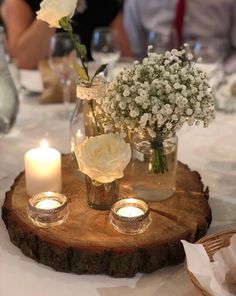 some white flowers and candles are sitting on a wood slice at a table with people in the background