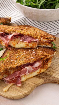 two sandwiches with meat, cheese and greens on a cutting board next to a bowl of salad