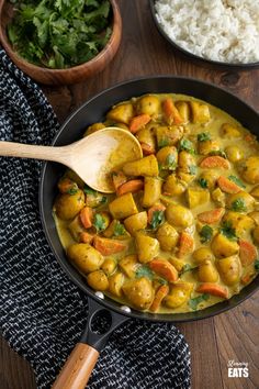 a pan filled with curry and vegetables on top of a wooden table next to rice