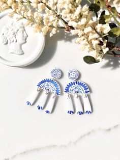 two white and blue earrings sitting on top of a table next to some dried flowers