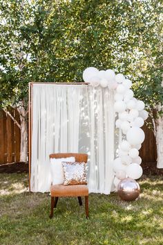 a chair and balloon arch in the grass at a backyard party with white balloons on it