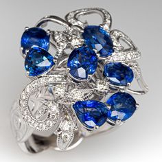 This wonderful cluster style ring is accented with five (5), prong set, oval mixed cut natural sapphires, one (1), prong set, pear mixed cut natural sapphire and one (1), prong set, triangular mixed cut natural sapphire. The sapphires are bordered with three (3), prong set, round brilliant cut diamonds and forty- nine (49), bead set, round brilliant/single cut diamonds, bordered with milgrain edging. Engraved details accent the shoulders of the shank. The ring measures 17.9mm at the top, rises 9.9mm above the finger, tapering to 3.3mm wide and 1.1mm thick at the base of the shank. This ring is currently a size 6.75. Round Brilliant Diamond Ring, Antique Cocktail Ring, Blue Sapphire Diamond, Bead Set, Dress Clothes, Diamond Cluster Ring, Natural Sapphire, Diamond Cluster, Brilliant Diamond