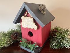 a red birdhouse sitting on top of a wooden table next to pine cones and evergreens